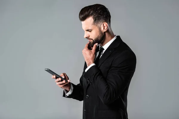 Vista lateral del hombre de negocios reflexivo en traje negro usando teléfono inteligente aislado en gris - foto de stock