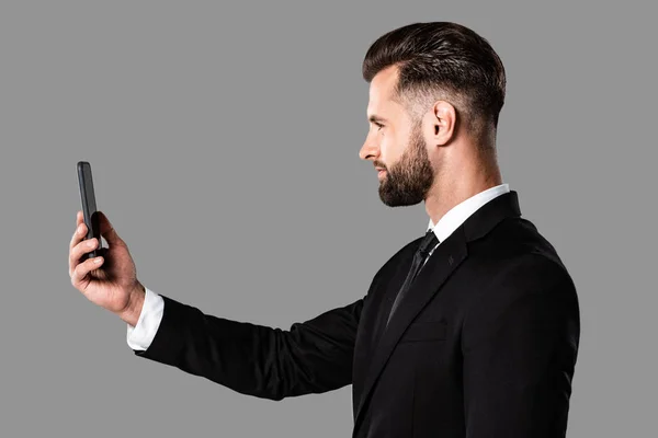 Вид сбоку красивого бизнесмена в черном костюме, делающего селфи на смартфоне, изолированном на сером — стоковое фото