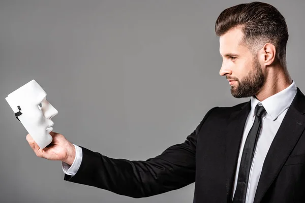 Side view of confident businessman in black suit looking at mask isolated on grey — Stock Photo