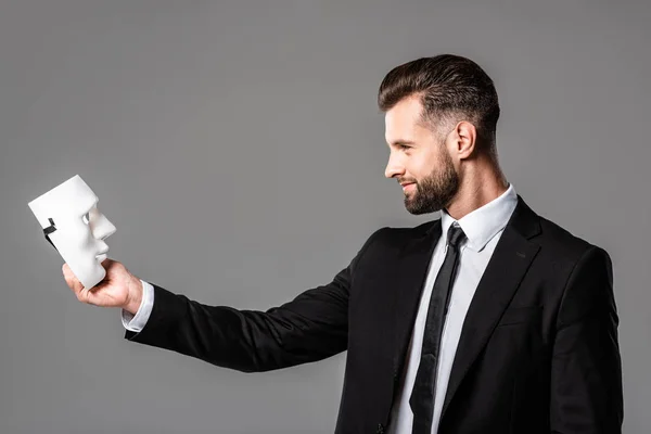 Side view of smiling businessman in black suit looking at mask isolated on grey — Stock Photo