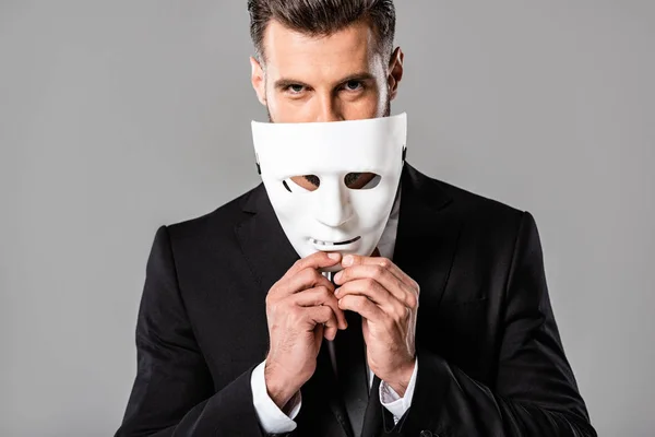 Astuto hombre de negocios guapo en traje negro quitándose la máscara blanca aislado en gris - foto de stock
