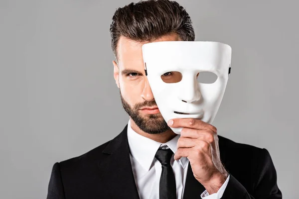 Hombre de negocios guapo serio en traje negro quitándose la máscara blanca aislado en gris - foto de stock