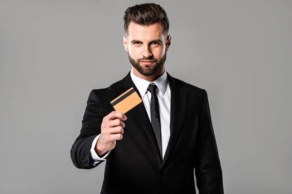 Bel homme d'affaires en costume noir présentant carte de crédit isolé sur gris — Photo de stock