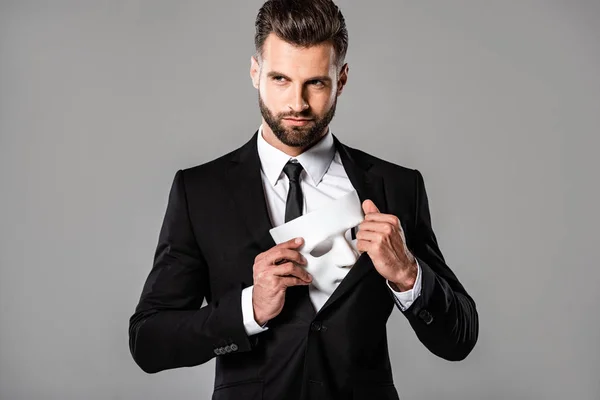 Хитромудрий бізнесмен в чорному костюмі ховає білу маску ізольовано на сірому — Stock Photo
