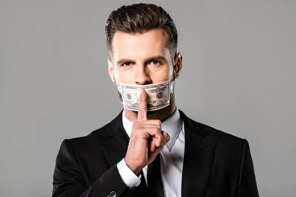 Businessman in black suit with dollar banknote on mouth showing hush gesture isolated on grey — Stock Photo