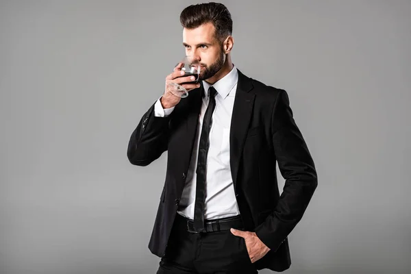 Handsome businessman in black suit drinking whiskey isolated on grey — Stock Photo