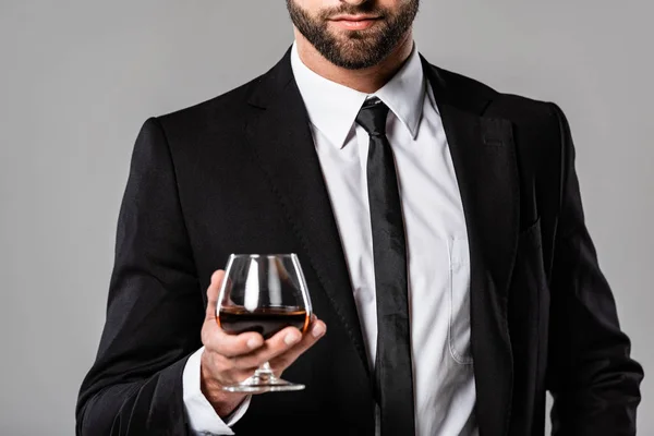 Vista recortada del hombre de negocios barbudo en traje negro sosteniendo vidrio con whisky aislado en gris - foto de stock