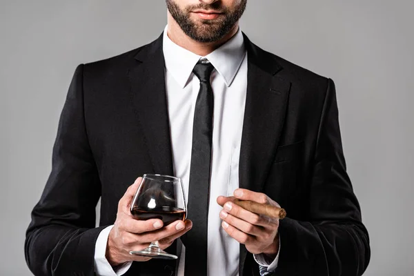 Vista cortada de empresário em terno preto segurando vidro com uísque e charuto isolado em cinza — Fotografia de Stock