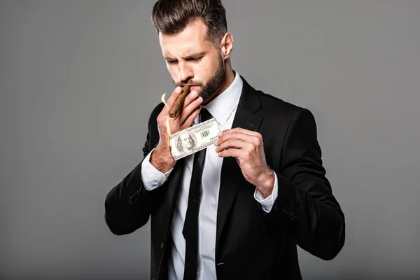 Rico exitoso hombre de negocios en traje negro iluminando cigarro de billete de dólar en llamas aislado en gris — Stock Photo