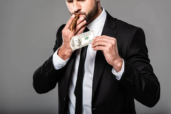 Vista recortada de rico empresario exitoso en traje negro iluminando cigarro de billete de dólar en llamas aislado en gris - foto de stock