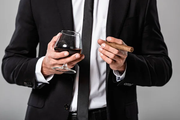 Vista parcial del hombre de negocios en traje negro sosteniendo vidrio con whisky y cigarro aislado en gris - foto de stock