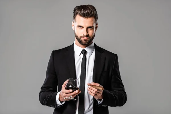 Exitoso hombre de negocios en traje negro sosteniendo vidrio con whisky y cigarro aislado en gris - foto de stock