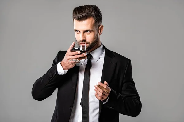 Guapo hombre de negocios en traje negro bebiendo whisky aislado en gris - foto de stock