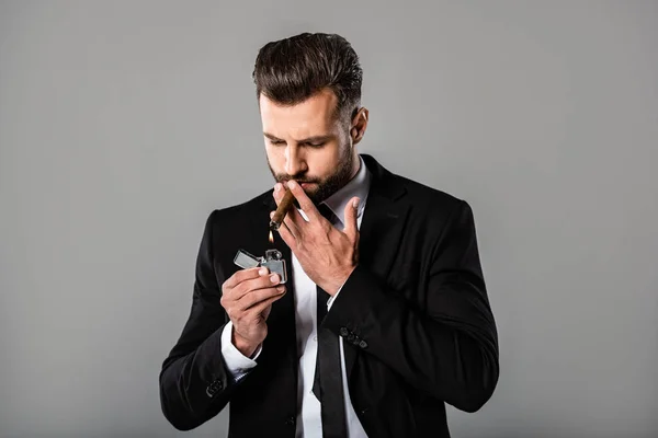 Успішний бізнесмен в чорному костюмі освітлює сигару ізольовано на сірому — стокове фото