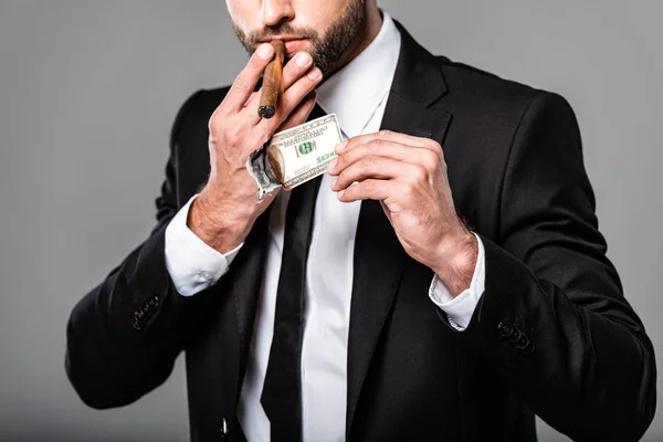 Vista parcial de rico empresario exitoso en traje negro iluminando cigarro de billete de dólar en llamas aislado en gris - foto de stock
