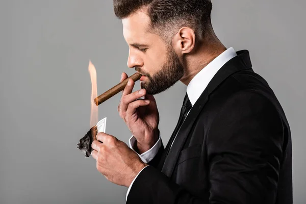 Side view of rich successful businessman lighting up cigar from burning dollar banknote isolated on grey — Stock Photo