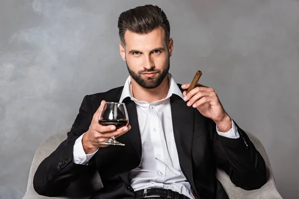 Empresário de sucesso em terno preto sentado em poltrona com charuto e uísque em fumaça — Fotografia de Stock
