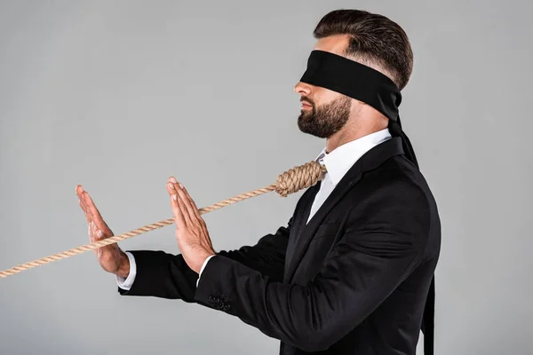 Side view of blindfolded businessman in black suit with noose on neck showing stop gesture isolated on grey — Stock Photo