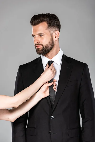 Mujer fijación corbata en exitoso hombre de negocios en traje negro aislado en gris - foto de stock