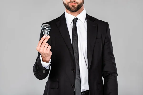 Vista recortada del exitoso hombre de negocios en traje negro sosteniendo bombilla aislada en gris - foto de stock