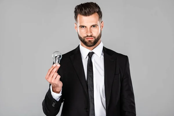 Exitoso hombre de negocios en traje negro sosteniendo bombilla aislada en gris - foto de stock