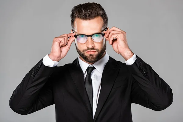 Exitoso hombre de negocios en traje negro tocando gafas aisladas en gris - foto de stock