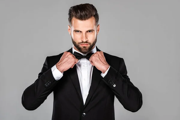Елегантний впевнений чоловік в чорному костюмі фіксує краватку ізольовано на сірому — стокове фото