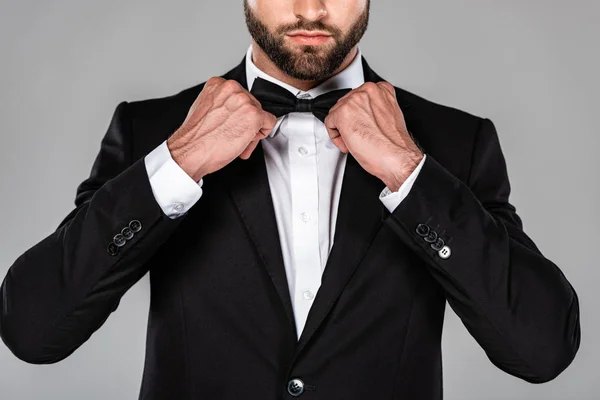 Vista recortada de hombre elegante en traje negro fijación pajarita aislado en gris - foto de stock