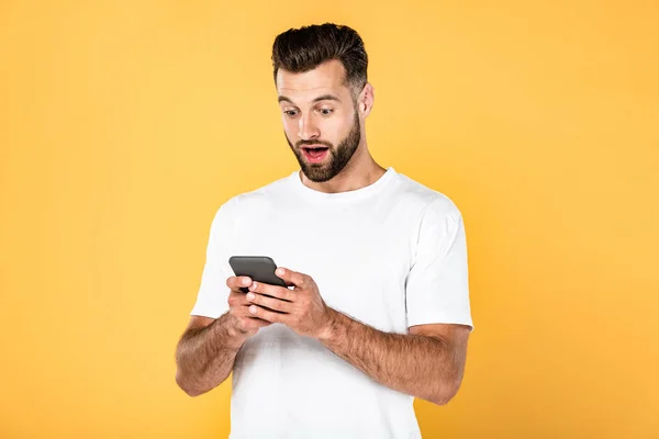 Excité bel homme en t-shirt blanc en utilisant smartphone isolé sur jaune — Photo de stock