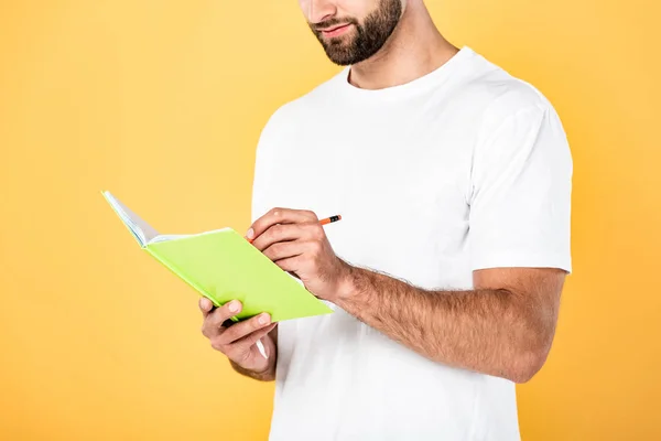 Vista recortada de hombre guapo en camiseta blanca escrito en cuaderno aislado en amarillo - foto de stock