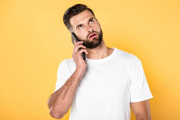 Bel homme réfléchi en t-shirt blanc parlant sur smartphone isolé sur jaune — Photo de stock