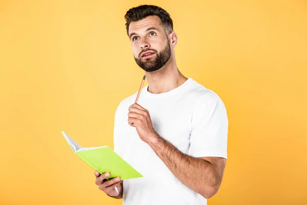 Homem bonito pensativo na t-shirt branca que segura o lápis e o caderno isolados no amarelo — Fotografia de Stock