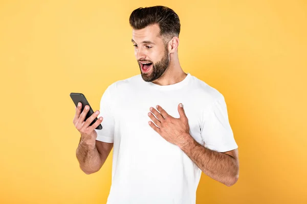 Hombre guapo excitado en camiseta blanca usando teléfono inteligente aislado en amarillo - foto de stock