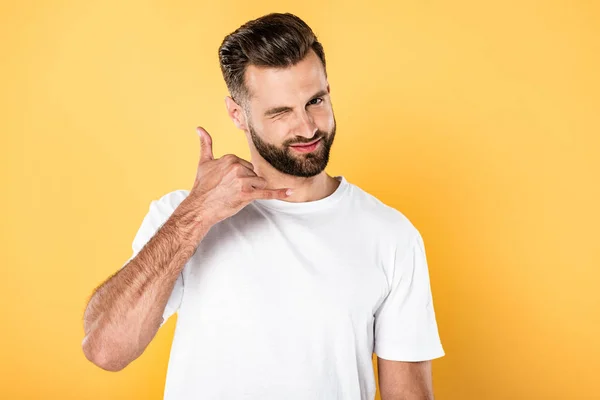 Augenzwinkernder gutaussehender Mann im weißen T-Shirt shawing rufen Sie mich Geste isoliert auf gelb — Stockfoto