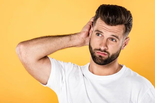 Hombre reflexivo en camiseta blanca arañando la cabeza aislado en amarillo - foto de stock