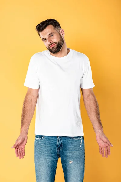 Homem bonito sorridente na t-shirt branca mostrando gesto de dubium isolado no amarelo — Fotografia de Stock