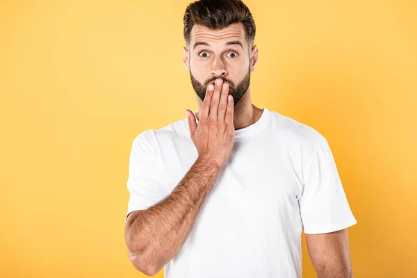 Shocked handsome man in white t-shirt with hand on mouth isolated on yellow — Stock Photo