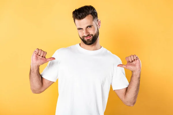 Улыбающийся красивый мужчина в белой футболке, указывающий пальцами на себя, изолированный на желтом — стоковое фото