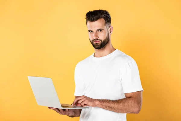 Homem bonito em t-shirt branca usando laptop isolado no amarelo — Fotografia de Stock