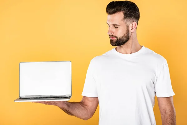 Красивий чоловік у білій футболці дивиться на ноутбук з порожнім екраном ізольовано на жовтому — стокове фото