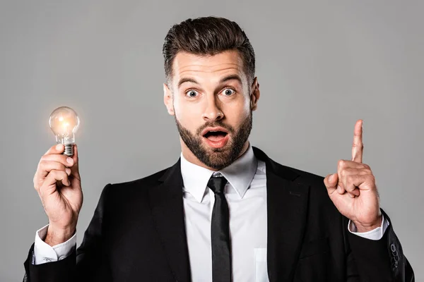 Surprised businessman in black suit holding glowing light bulb and showing idea gesture isolated on grey — Stock Photo