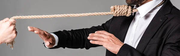 Vista recortada del hombre de negocios en traje negro con soga en el cuello mostrando gesto de parada al asesino con cuerda aislada en gris, tiro panorámico - foto de stock