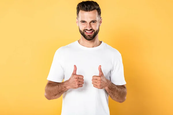 Hombre guapo sonriente en camiseta blanca mirando a la cámara y mostrando los pulgares hacia arriba aislado en amarillo - foto de stock