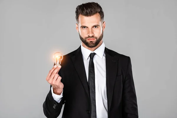 Hombre de negocios en traje negro sosteniendo bombilla brillante aislado en gris - foto de stock