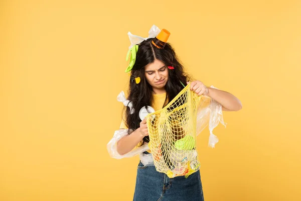 Mujer joven confundida con la basura en la bolsa de la celebración del pelo con la basura aislada en amarillo - foto de stock