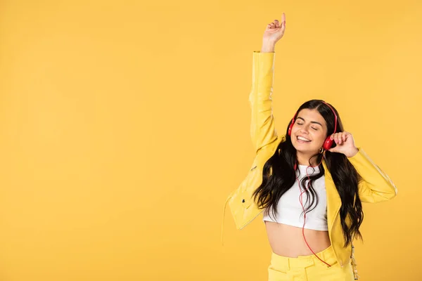 Alegre chica bailando y escuchando música con auriculares, aislado en amarillo - foto de stock