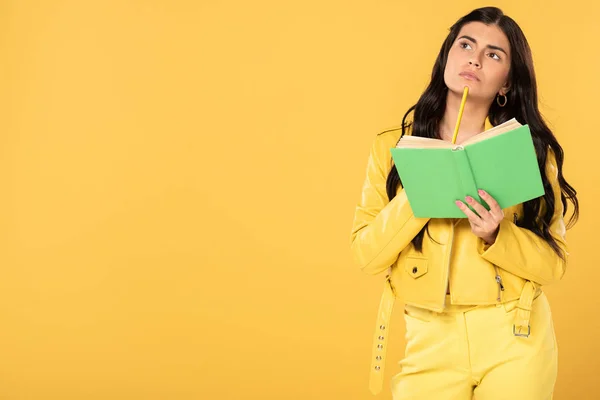 Chica pensativa sosteniendo lápiz y libro, aislado en amarillo - foto de stock