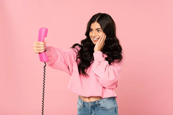 Приваблива усміхнена дівчина дивиться на ретро телефон ізольовано на рожевому — стокове фото