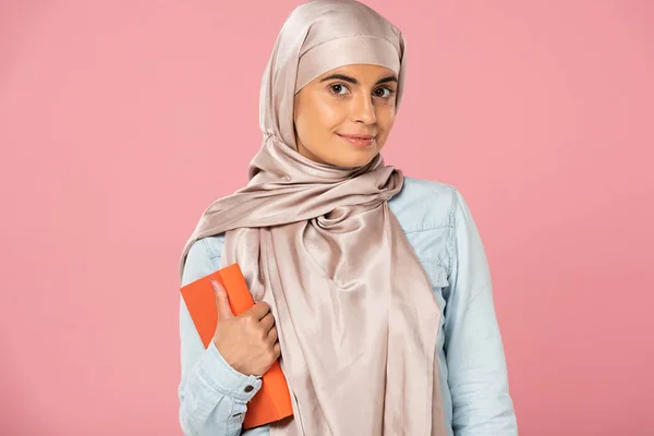 Attractive muslim student in hijab holding book isolated on pink — Stock Photo