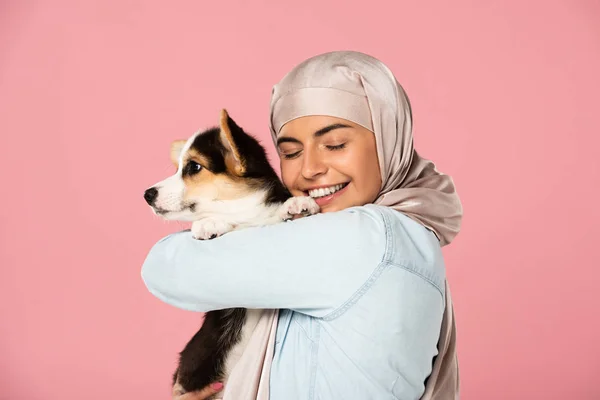 Happy arabic woman in hijab holding Welsh Corgi puppy, isolated on pink — Stock Photo